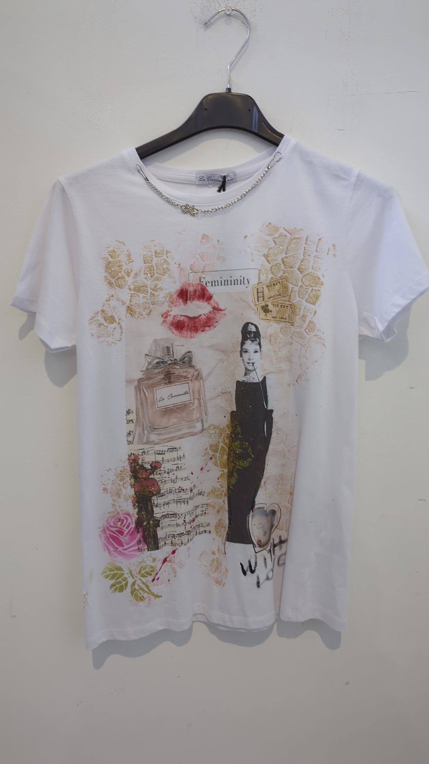 Romantica, chic e di grande impatto visivo: questa t-shirt bianca da donna celebra la femminilità con una stampa artistica ispirata all’eleganza retrò. Dettagli dorati glitterati, labbra rosse, note musicali, rosa stampata e un profumo "La Coccinella" arricchiscono il capo, rendendolo perfetto per look raffinati e originali.