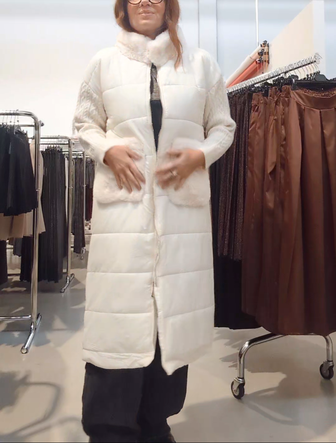 Donna che indossa un piumino lungo bianco con collo in pelliccia ecologica, maniche in maglia e tasche morbide, in negozio di abbigliamento.